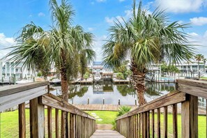 A wooden deck leading to a water feature with palm trees, a boat dock, and modern buildings in the background. - Joy by the Sea - Large Home w/ Harbor Views - Pet Friendly & Private Dock (Destin)