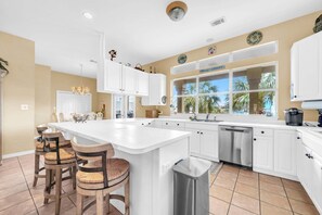 A spacious kitchen with white cabinetry, a large island with bar stools, and a view of palm trees through the windows. - Joy by the Sea - Large Home w/ Harbor Views - Pet Friendly & Private Dock (Destin)