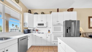 Une cuisine moderne avec des meubles blancs, un réfrigérateur en acier inoxydable et une fenêtre donnant sur des palmiers.
