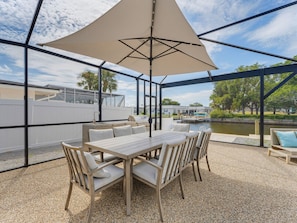 Eine Terrasse im Freien mit Tisch und Stühlen, einem großen Sonnenschirm und Blick auf ein Gewässer sowie Gebäude im Hintergrund.