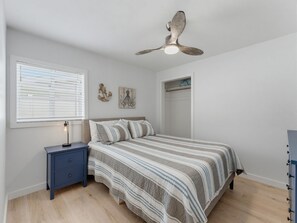 Ein Schlafzimmer mit einem Bett, einem Nachttisch, einem Deckenventilator, einem Fenster mit Jalousien und einer Wand mit zwei Kunstwerken.