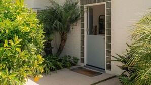 A modern house with a white exterior, a covered porch, and a variety of greenery including palm trees and shrubs. - A Mesa Gem with Chic Coastal Vibes, Lush Outdoor Space, Detached Guest Studio & Hot Tub! (Santa Barbara)