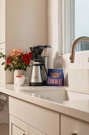 A kitchen counter with a coffee maker, a mug, a vase with flowers, and a bag of coffee beans. - A Mesa Gem with Chic Coastal Vibes, Lush Outdoor Space, Detached Guest Studio & Hot Tub! (Santa Barbara)