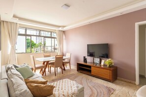 A living room with a sofa, a dining table with chairs, a TV on a wooden stand, and a view of buildings through the window. - Shopping Next Door | 3BR + Parking in Barra (Salvador)