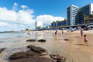 A beach with people, a lighthouse, and buildings in the background. - Shopping Next Door | 3BR + Parking in Barra (Salvador)