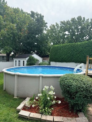 A backyard with a swimming pool, surrounded by a white fence, trees, and a hedge.