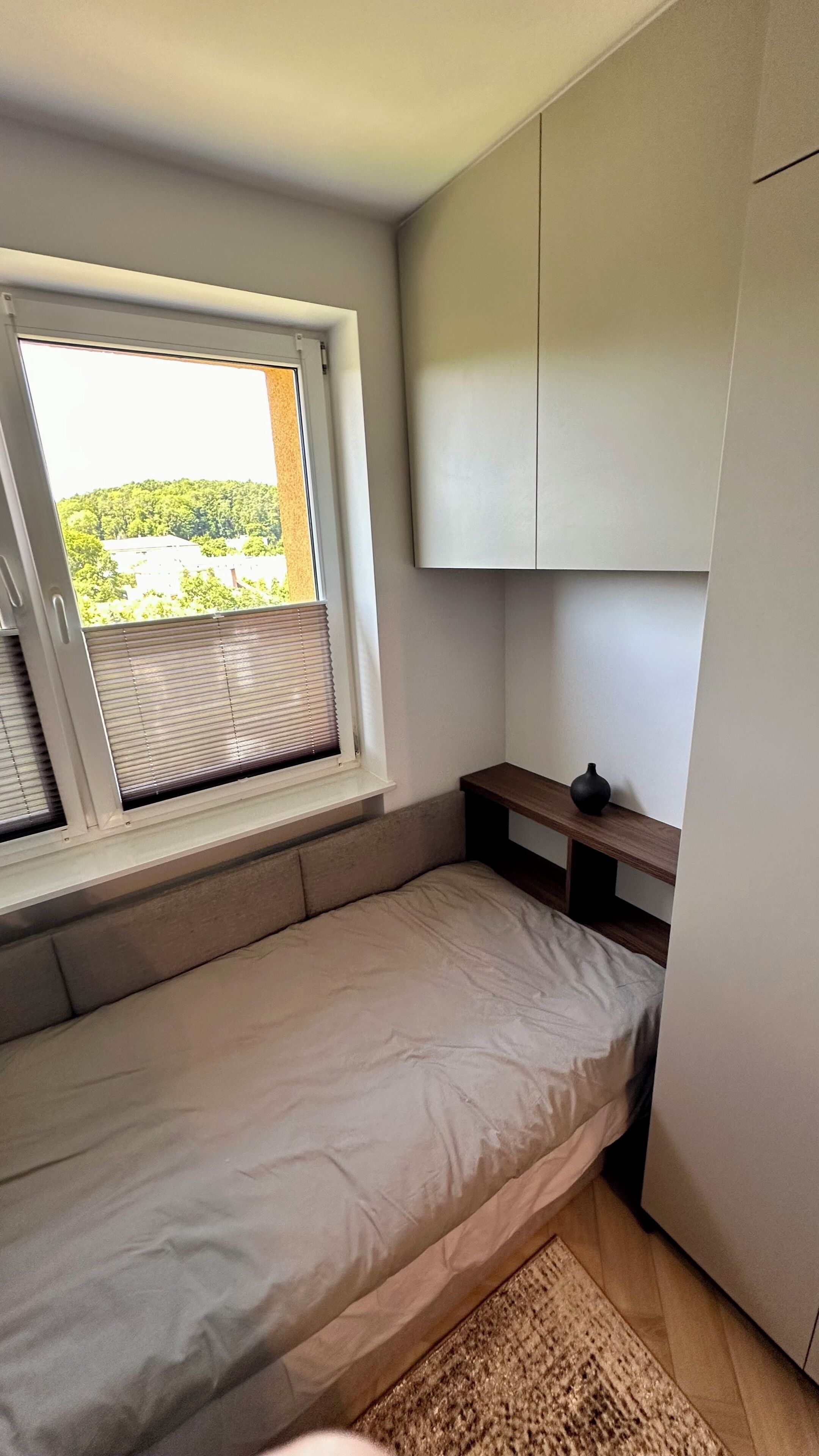 A small bedroom with a single bed, a window with blinds, a built-in wardrobe, and a small table.
