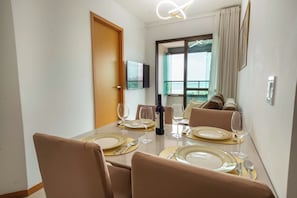 A dining area with a table set for four, a television mounted on the wall, and a view of the sea through a large window. - Ocean View at Corsário Beach | Pool & A/C (Salvador)