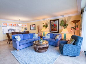 A living room with a blue sofa set, a coffee table, and a patterned armchair. A kitchen is visible in the background. - Sanibel Harbour Resort -Harbour Tower 913: Enjoy the most spectacular waterfront sunsets. (Fort Myers)