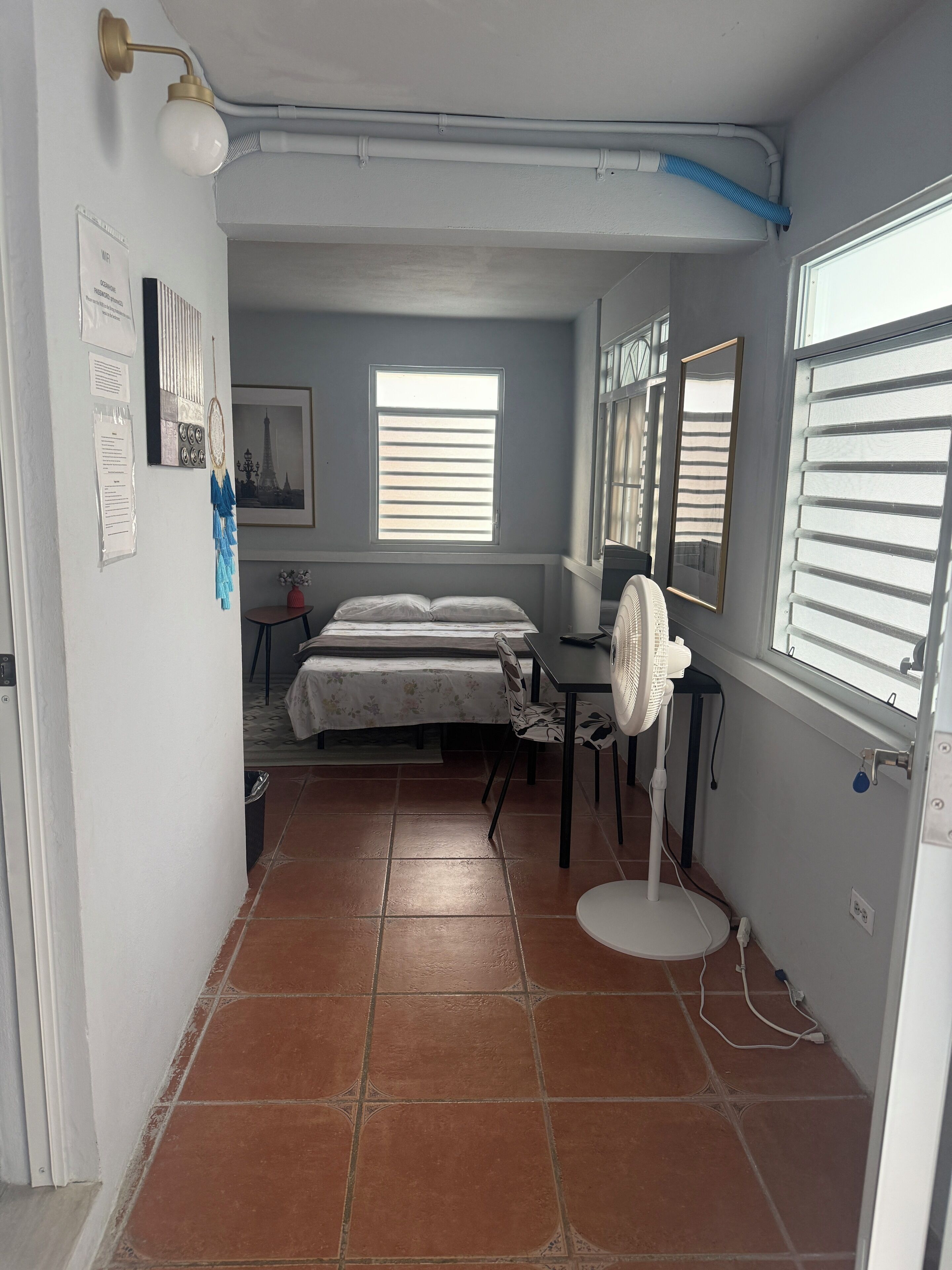 Ein kleiner, schmaler Raum mit einem Bett, einem Schreibtisch und einem Stuhl. Zwei Fenster mit Jalousien, ein Wandventilator und eine sichtbare Steckdose.