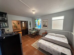 A compact bedroom with a bed, a desk, and a chair, a small table with a vase, and a window with blinds.