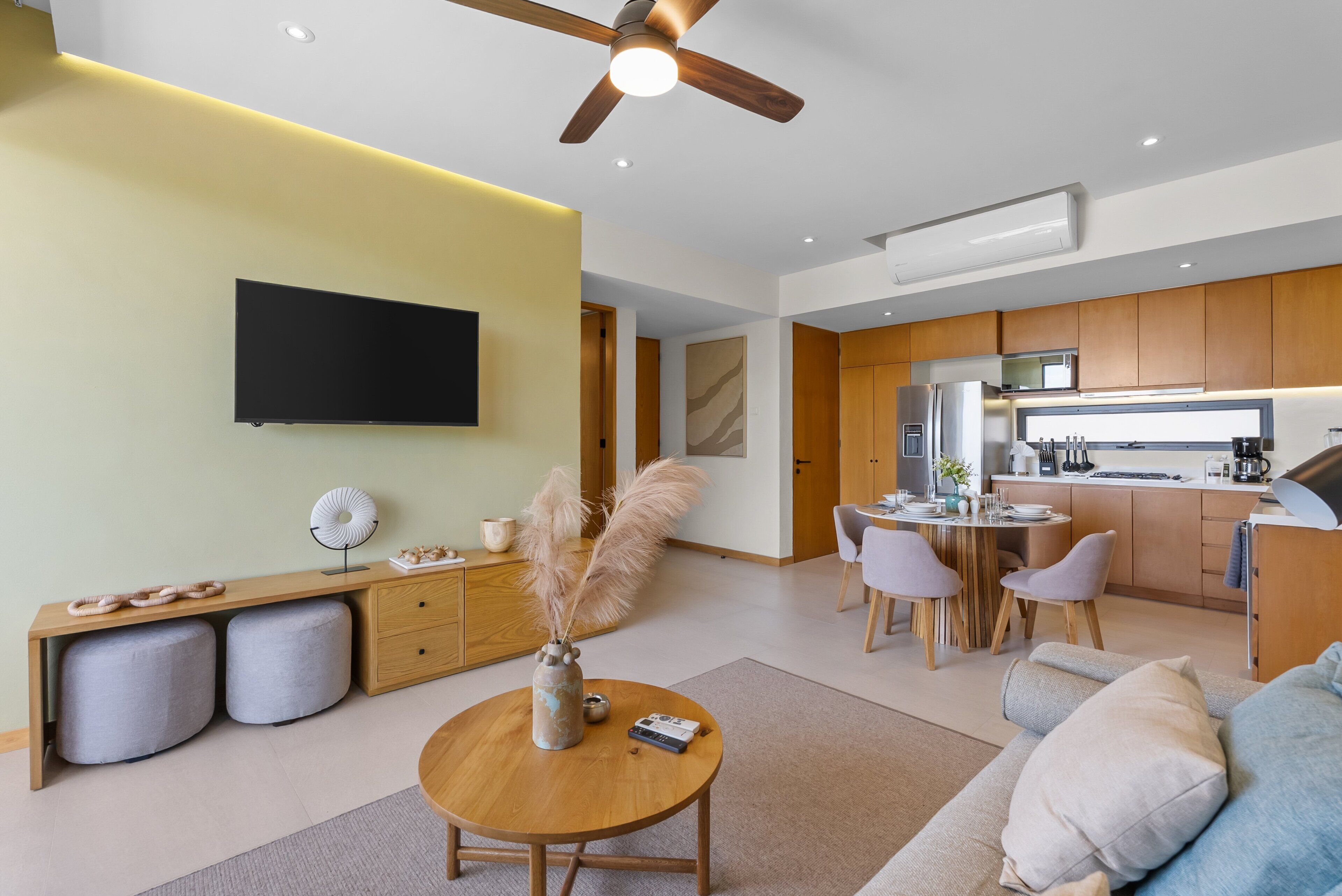 A modern living room with a flat-screen TV, a wooden dining table, and a kitchen area with wooden cabinets and a built-in oven.