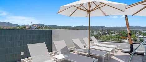A rooftop terrace with lounge chairs and umbrellas, overlooking a cityscape and hills.