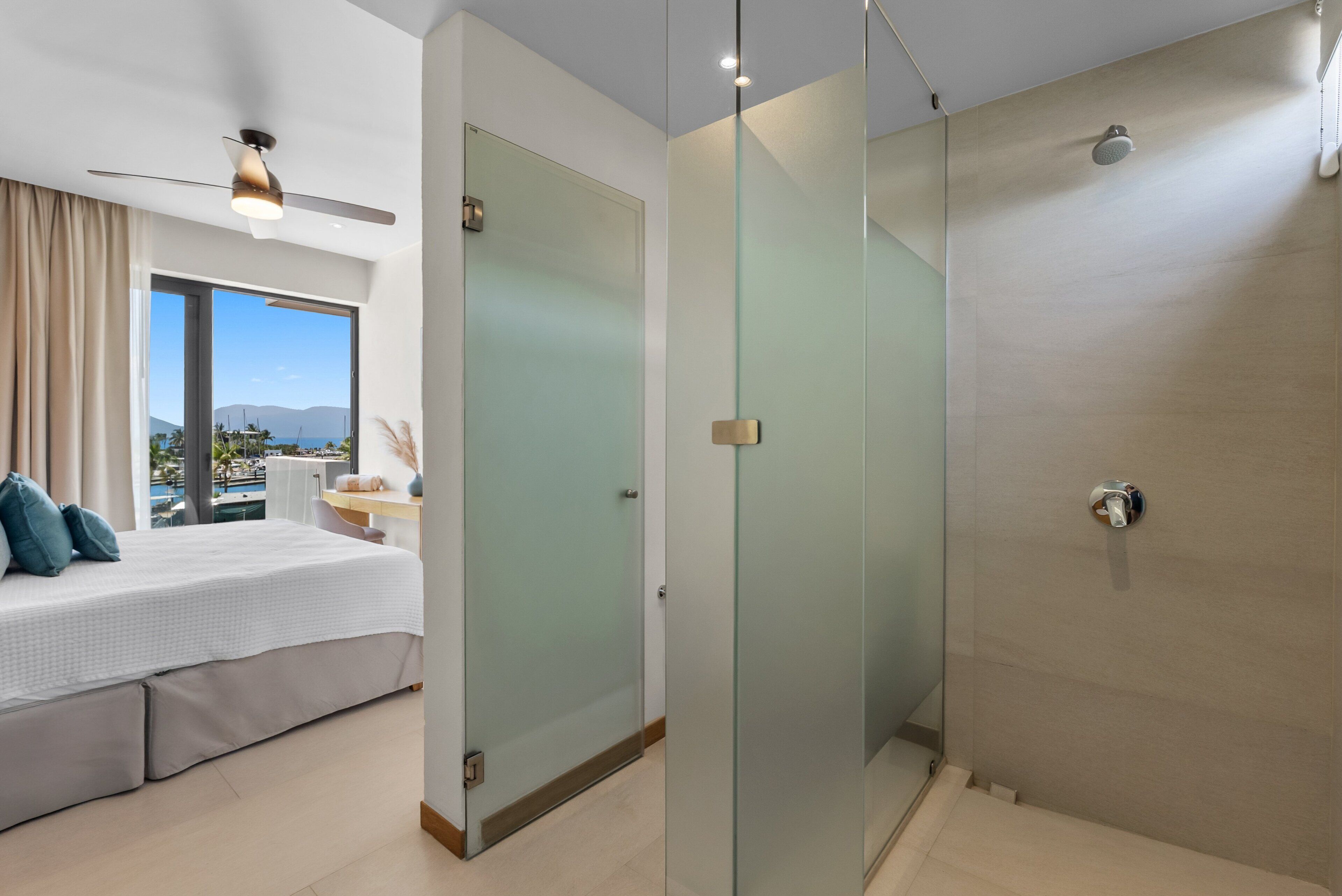 A modern hotel room with a bed, a desk, a glass shower enclosure, and a view of the outdoors.