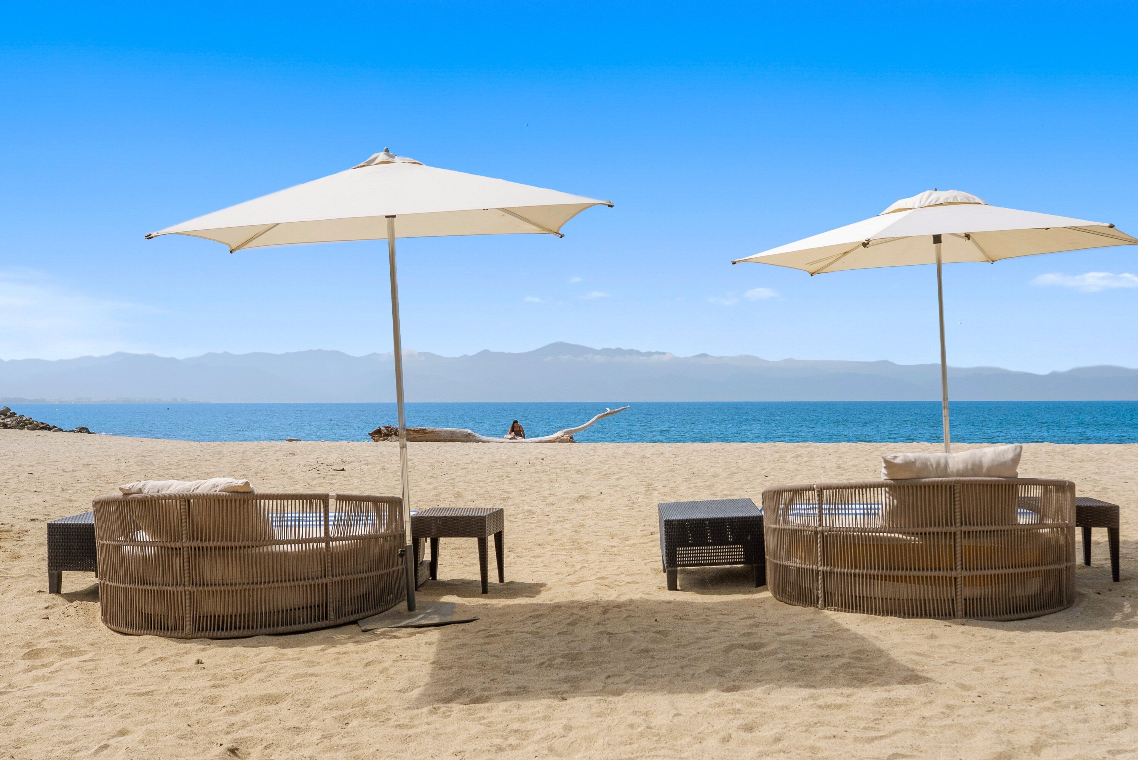 Een strand met twee rieten ligstoelen onder parasols, een persoon die ontspant op het zand, en een uitzicht op de zee en verre bergen.