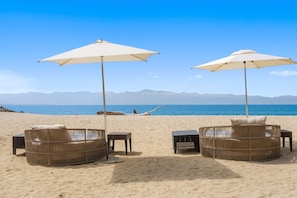 Una playa con dos sillones de mimbre bajo sombrillas, una persona relajándose en la arena, y vistas al mar y montañas distantes.