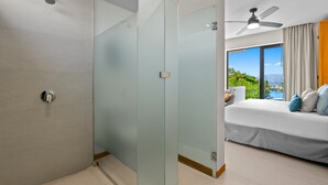Une chambre d’hôtel moderne avec une cloison en verre, un lit avec des oreillers, un ventilateur de plafond et une vue sur la verdure et un plan d’eau.