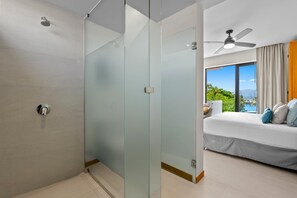 A modern hotel room with a glass partition, a bed with pillows, a ceiling fan, and a view of greenery and a body of water. - 2BR, 2BA Alik A5 condo in La Cruz with pool, gym, balcony, AC, WiFi. (Bahia de Banderas)