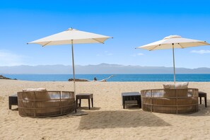 Ein Strand mit zwei Korbmöbel-Liegen unter Sonnenschirmen, eine Person entspannt sich im Sand, und es gibt einen Blick auf das Meer und ferne Berge.