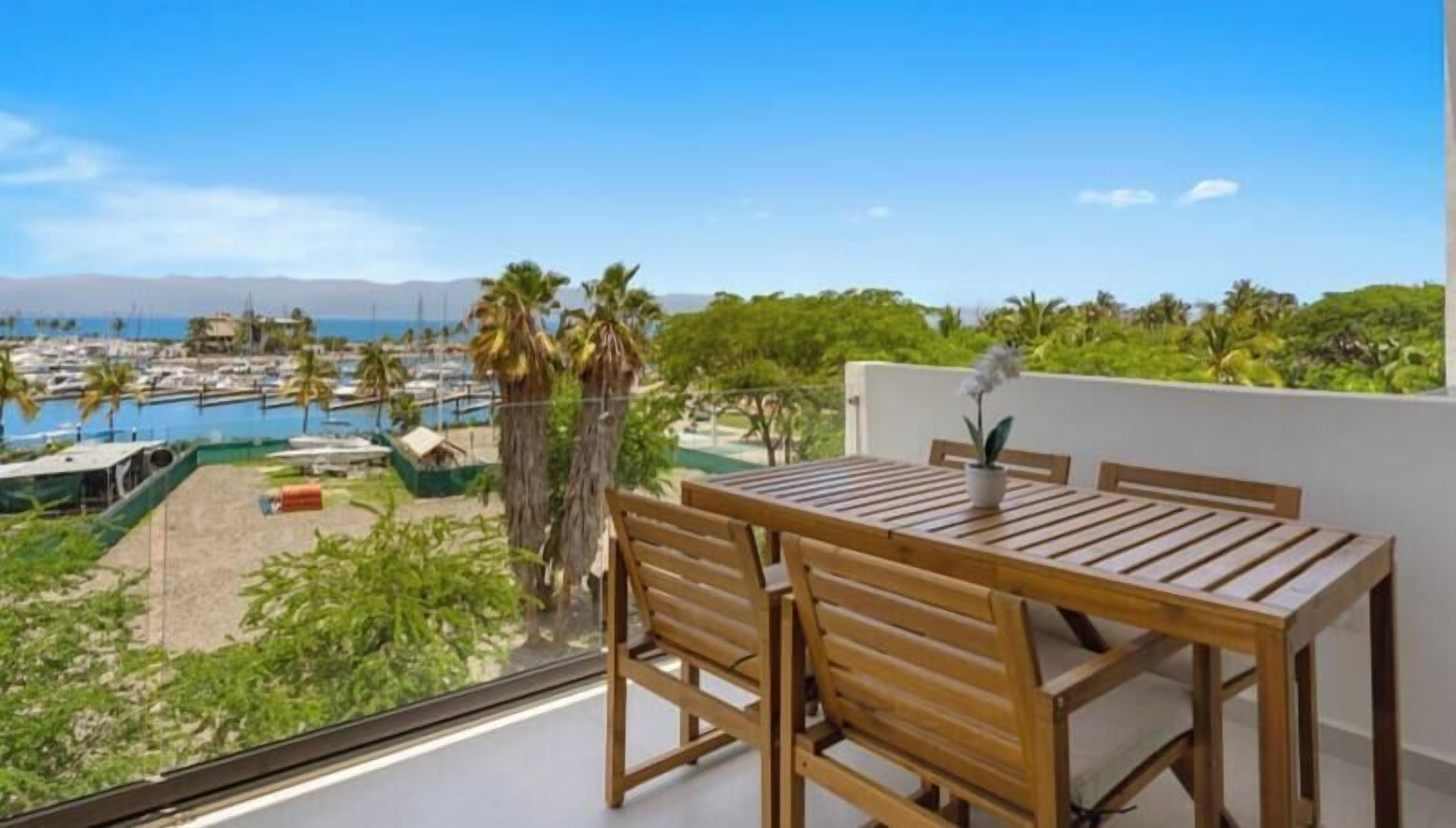 A wooden table and chairs on a balcony overlooking a marina with boats and palm trees.