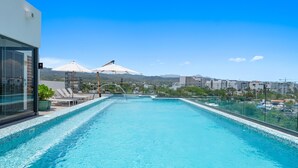 Uma piscina infinita no terraço, com águas azuis cristalinas, rodeada por espreguiçadeiras e guarda-sóis, com vista para uma paisagem urbana e montanhas ao longe.