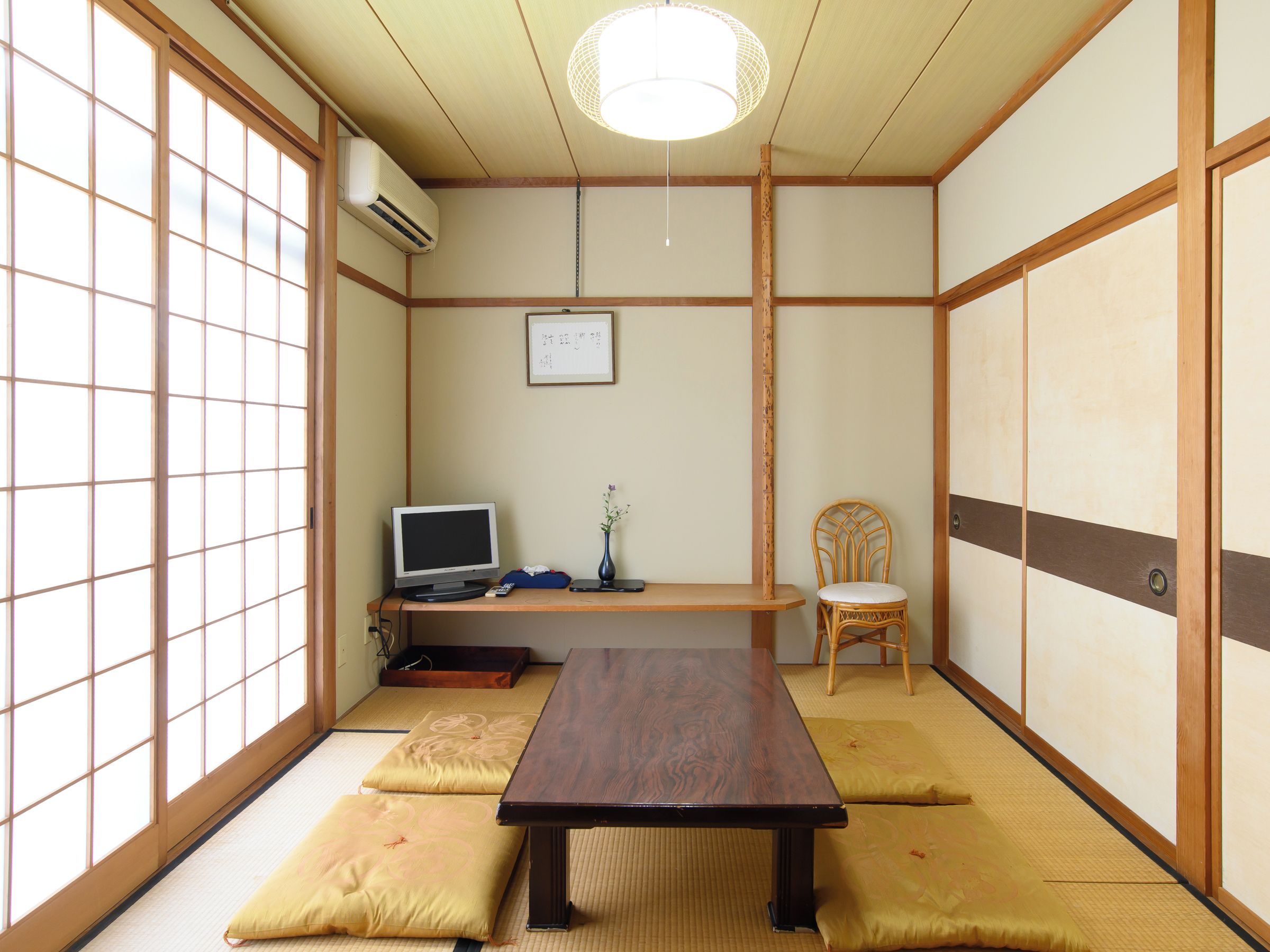 Una habitación tradicional japonesa con suelo de tatami, una mesa baja, una silla, un televisor y una lámpara colgante.