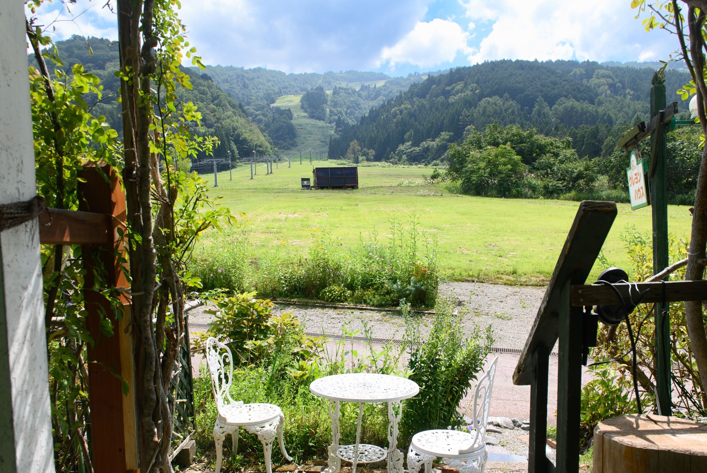 A view from a window overlooking a green field with a building in the distance. There are two white chairs and a small table in the foreground.