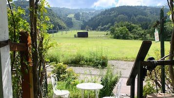 A view from a window overlooking a green field with a building in the distance. There are two white chairs and a small table in the foreground.