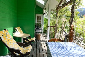 Una pared exterior verde, una terraza de madera con mesa y sillas, y una vista de un jardín con árboles y una montaña al fondo.