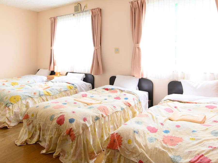 A hotel room with three beds, each with floral bedding, a bedside table with a lamp, and a window with curtains.