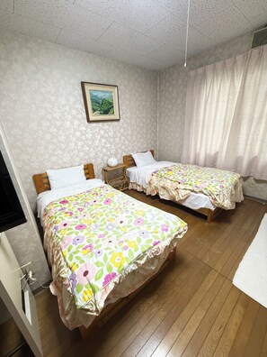 Quarto de hotel com duas camas, uma pequena mesa de cabeceira, televisão fixada na parede e uma janela com cortinas.