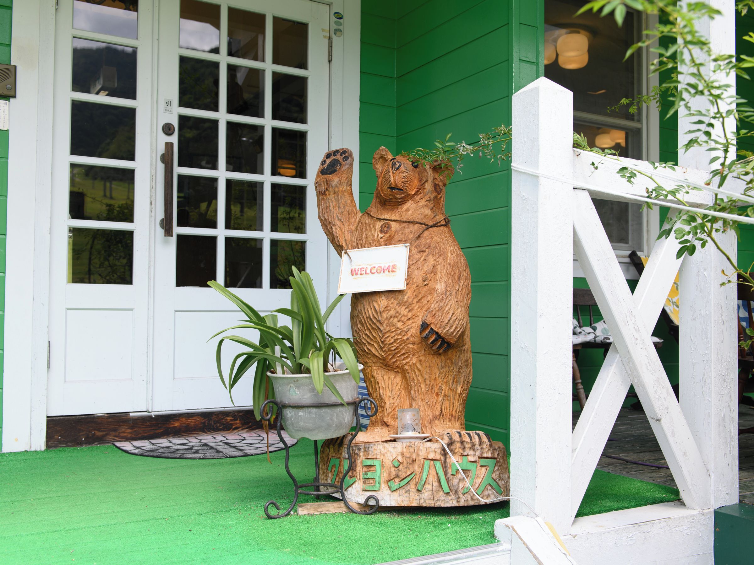 Una escultura de madera de un oso con un letrero que dice 'WELCOME', una planta en maceta y una base de madera con caracteres japoneses.