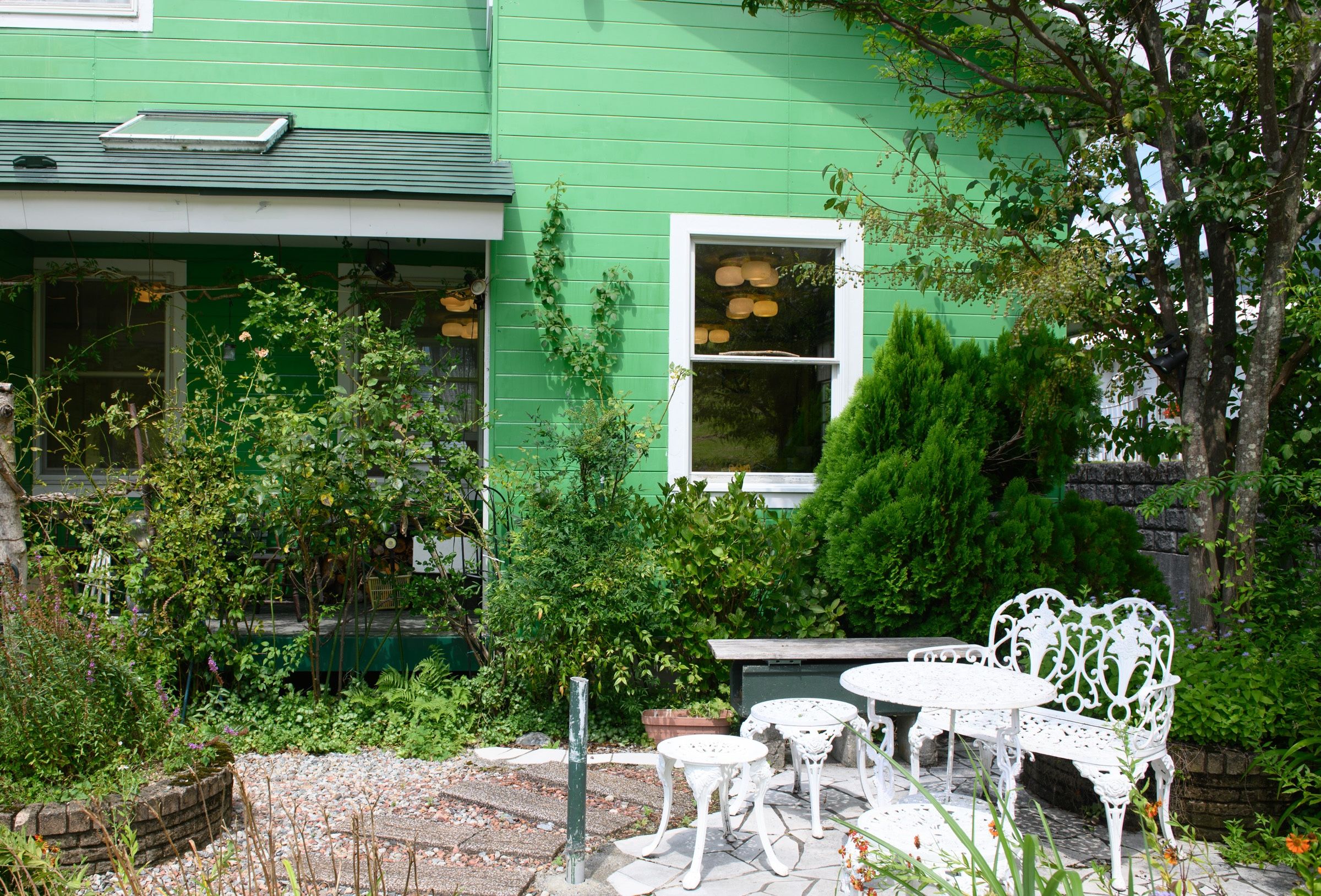 A green house with a garden, outdoor seating, and a window with hanging lights.