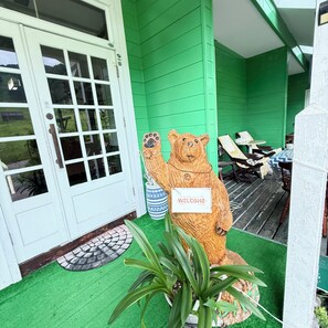 Fachada verde com porta branca, estátua de urso com placa de 'WELCOME', móveis de exterior e vaso de planta.