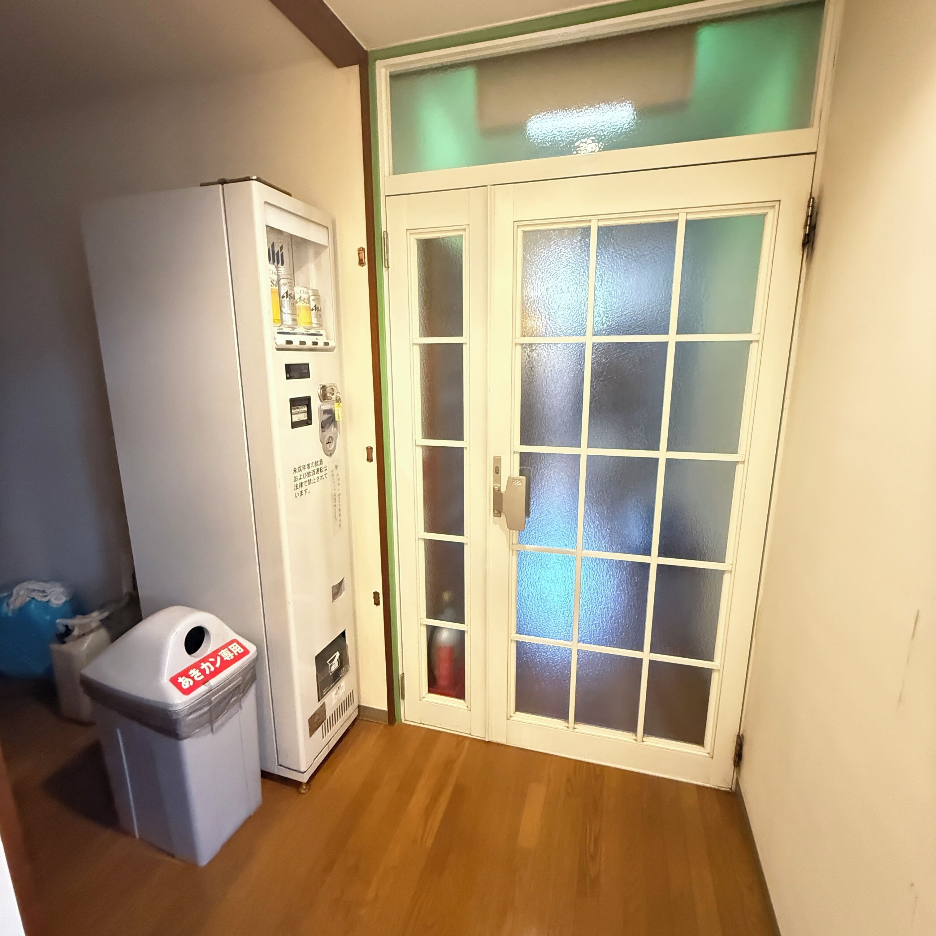 Una habitación con una máquina expendedora blanca, un cubo de basura y una puerta de cristal con un panel esmerilado.
