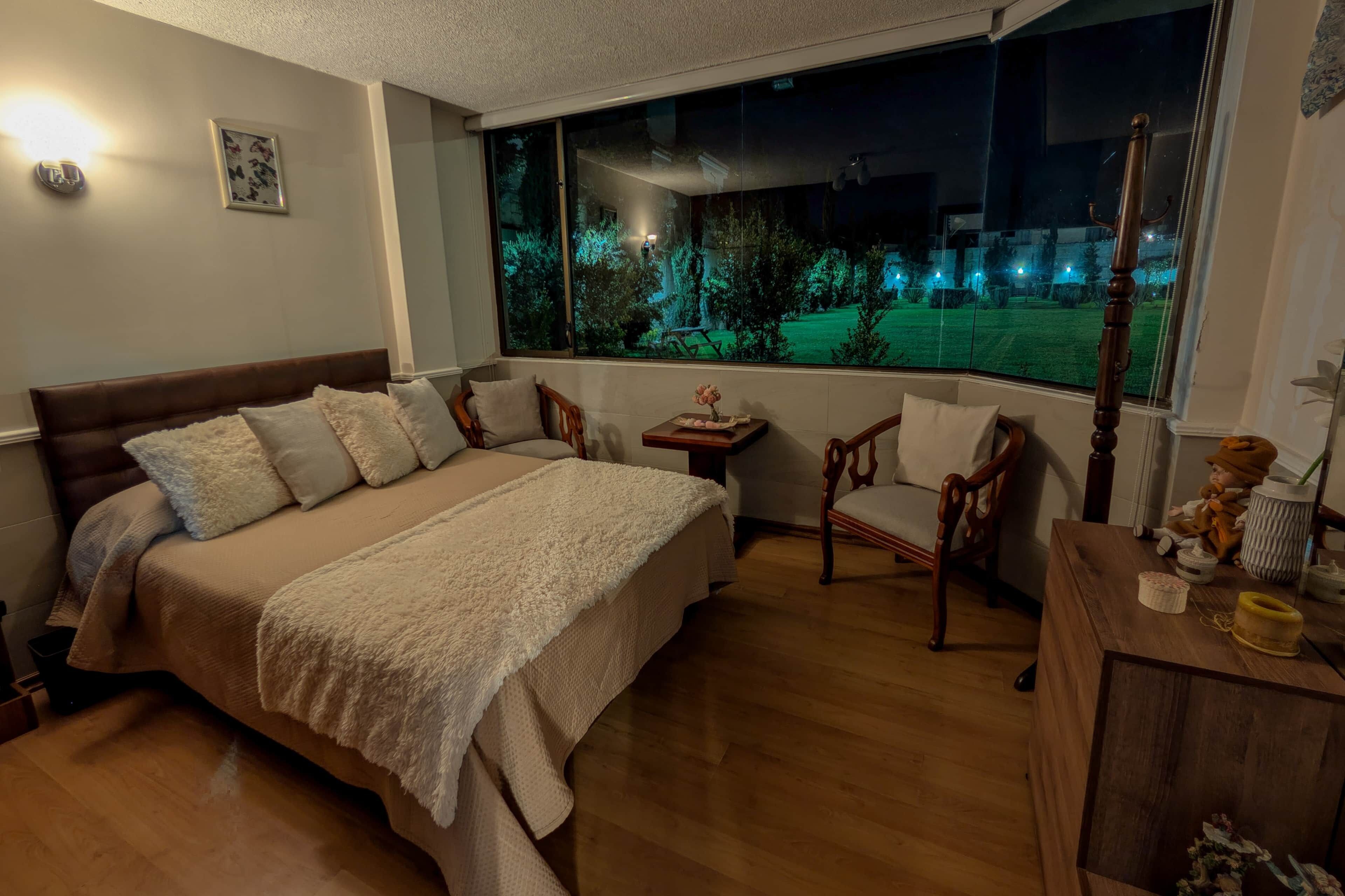 A bedroom with a large bed, a chair, a small table, and a window with a view of a garden at night.