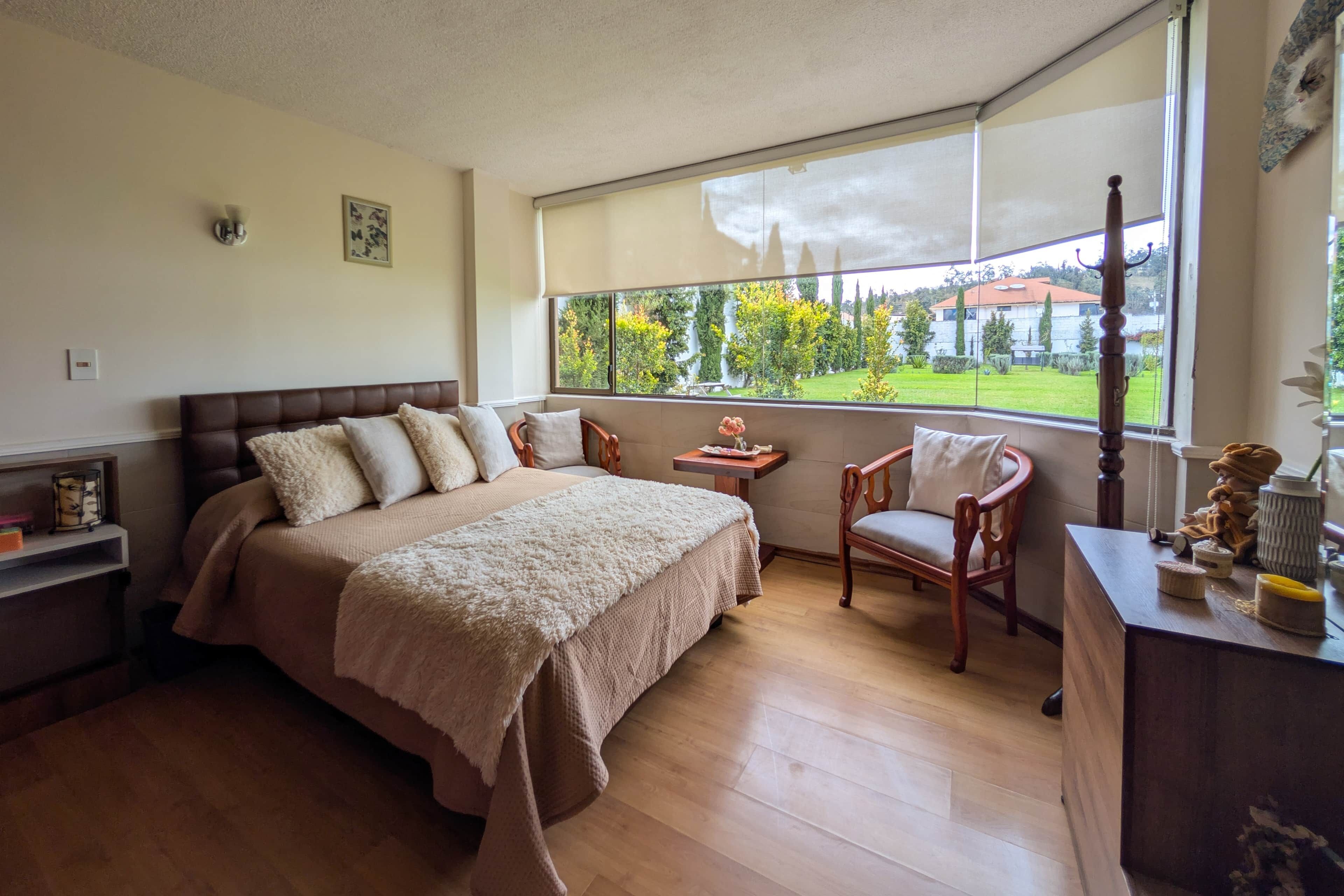 A bedroom with a large bed, a wooden chair, a small table, and a view of a garden through a window.