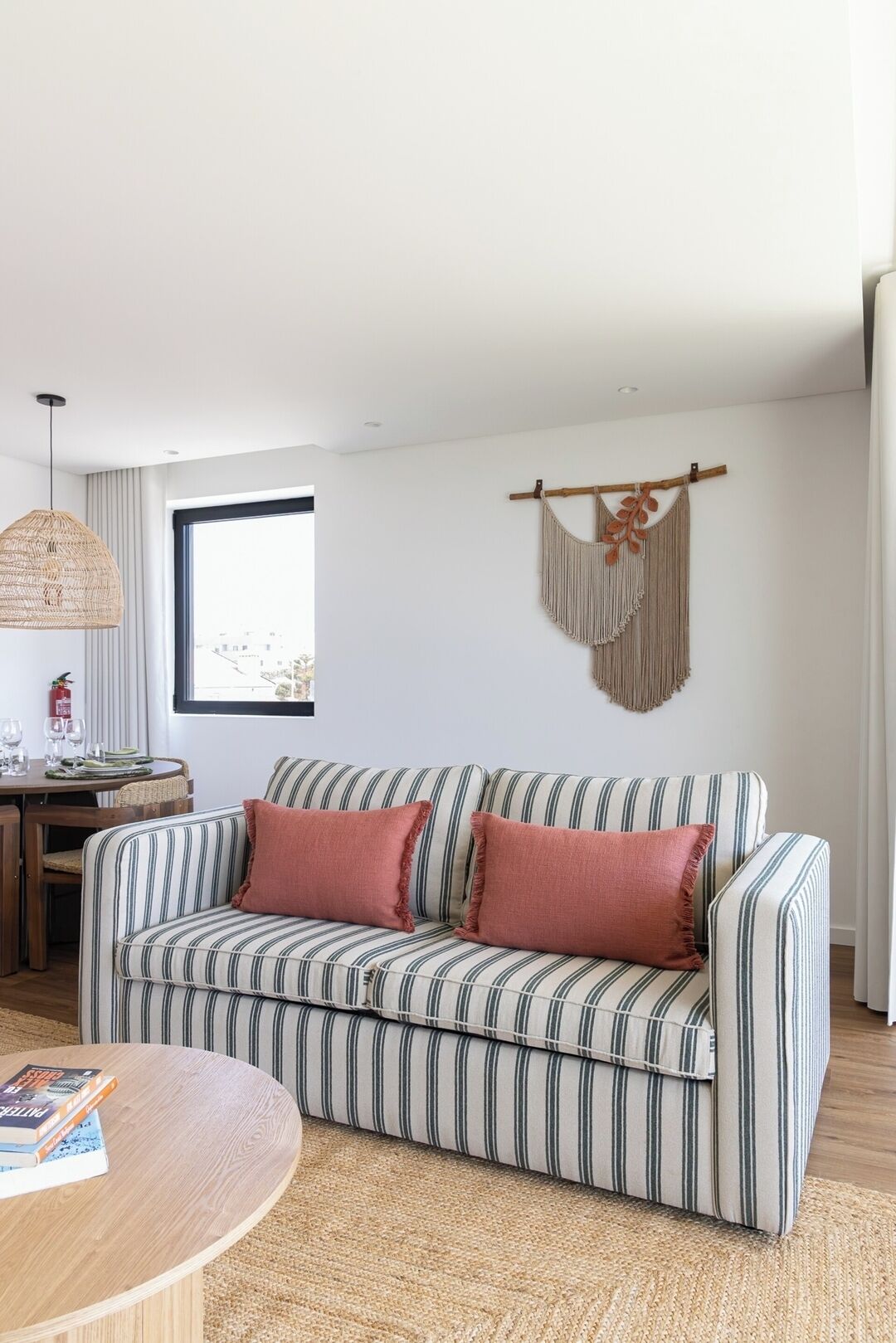 Un sofá a rayas con dos almohadones, una mesa redonda de madera con libros, y un tapiz de macramé en la pared.