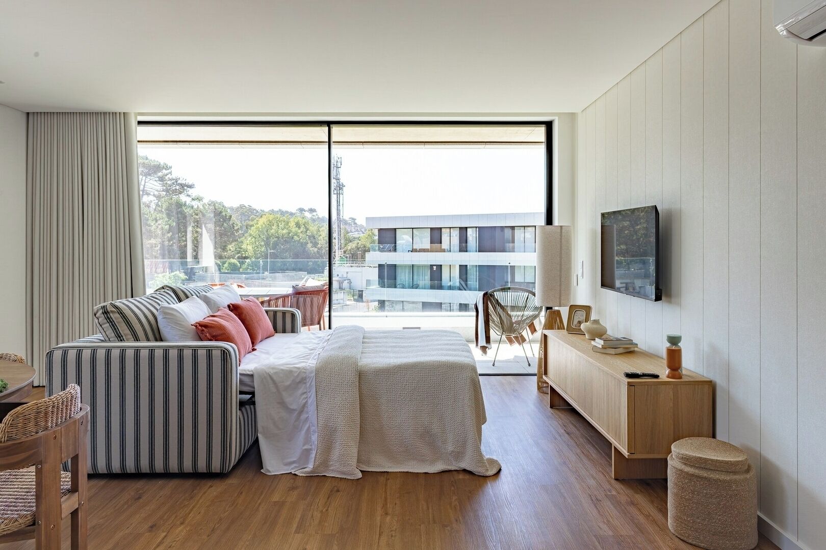 A modern bedroom with a large window, a bed, a sofa, a TV, and a wooden floor.