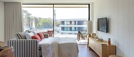 A modern bedroom with a large window, a bed, a sofa, a TV, and a wooden floor.