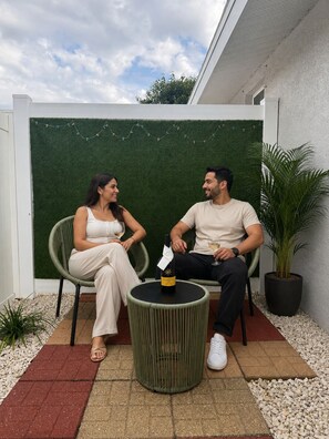 Zwei Personen sitzen an einem runden Tisch mit einer Weinflasche, auf einer Terrasse im Freien mit einer grünen Wand und Blumentöpfen im Hintergrund.
