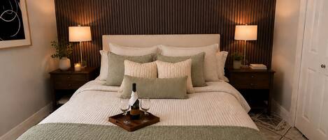 A hotel room with a large bed, two bedside tables with lamps, a framed picture on the wall, and a tray with a bottle and glasses on the bed.