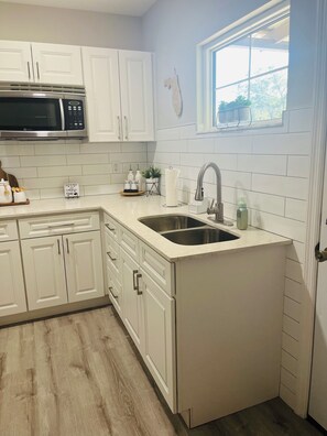 A kitchen with white cabinets, a stainless steel sink, a microwave, and a window with a view. - Little Piece of Paradise (Tampa)