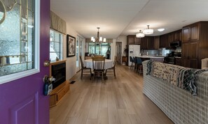Eine moderne Küche mit Holzschränken, ein Essbereich mit weißem Tisch und Stühlen sowie ein Wohnzimmer mit gemustertem Sofa.