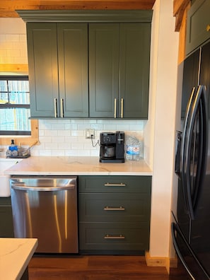 A kitchen with dark green cabinets, a stainless steel dishwasher, a coffee maker, and a refrigerator.