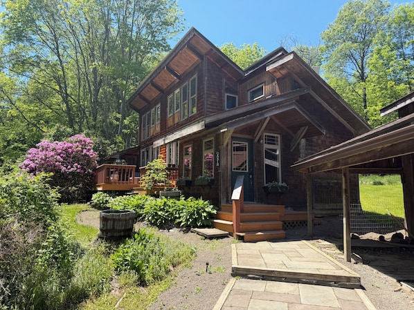 A two-story wooden cabin with a porch, surrounded by lush greenery and a well-maintained garden.