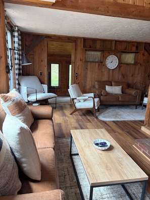 A cozy cabin interior with a wooden floor, a sofa, two armchairs, a coffee table, and a clock on the wall.