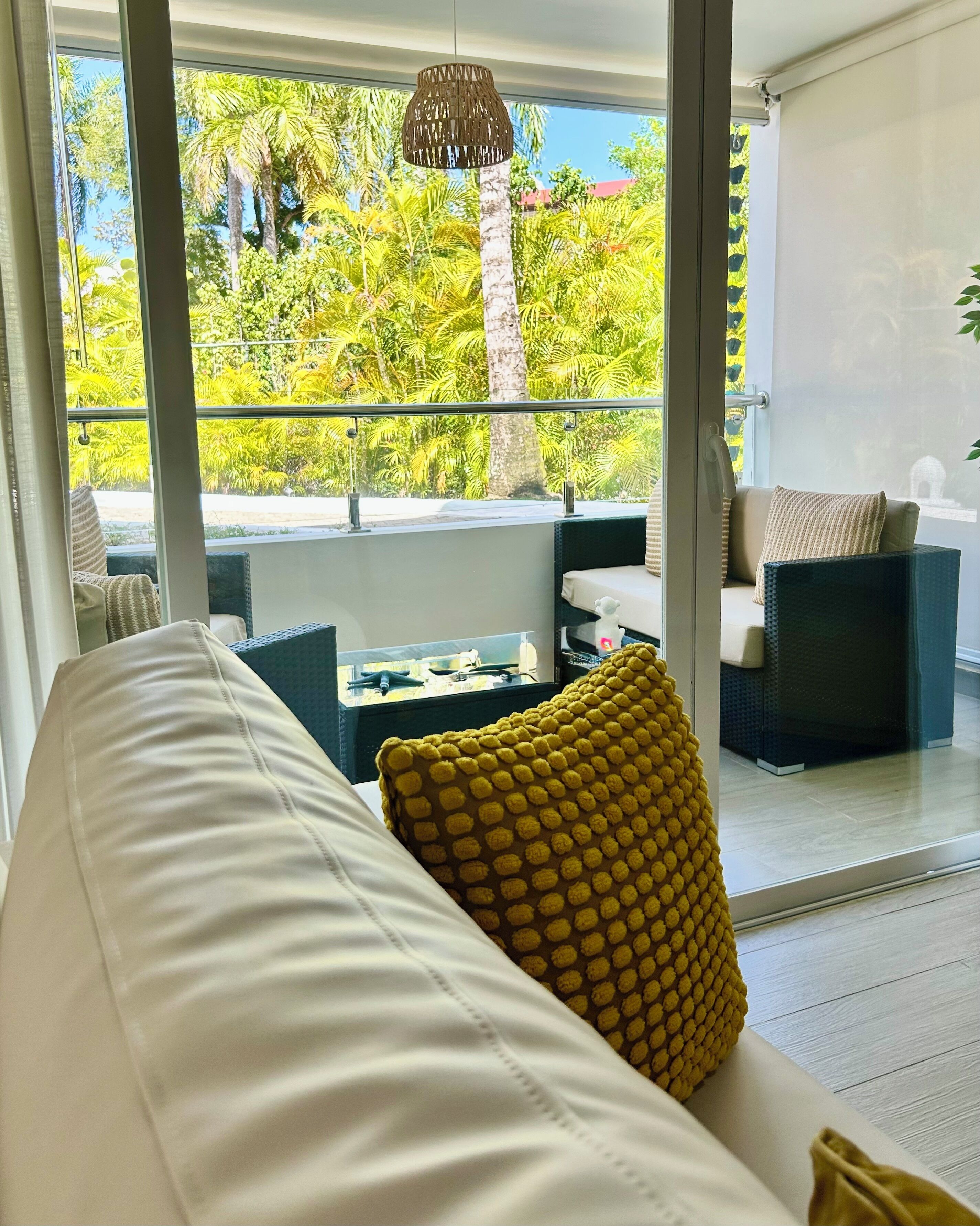 A room with a white sofa, a yellow patterned pillow, and a view of palm trees through large windows.