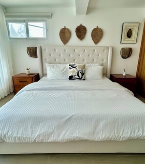 A large bed with a tufted headboard, white bedding, and decorative wall hangings. - 2BR Condo - Walk to Beach & Golf (Puerto Plata)