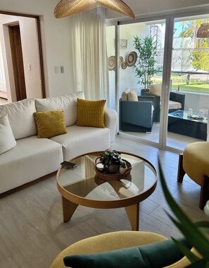 A modern living room with a sofa, a round coffee table, and a view of a garden through large glass doors. - 2BR Condo - Walk to Beach & Golf (Puerto Plata)
